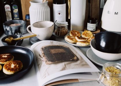 One portion of homemade cheese pancakes in frying pan and another on plate locating near opened stylish magazine among kitchen utensils and appliances in bright kitchen at home